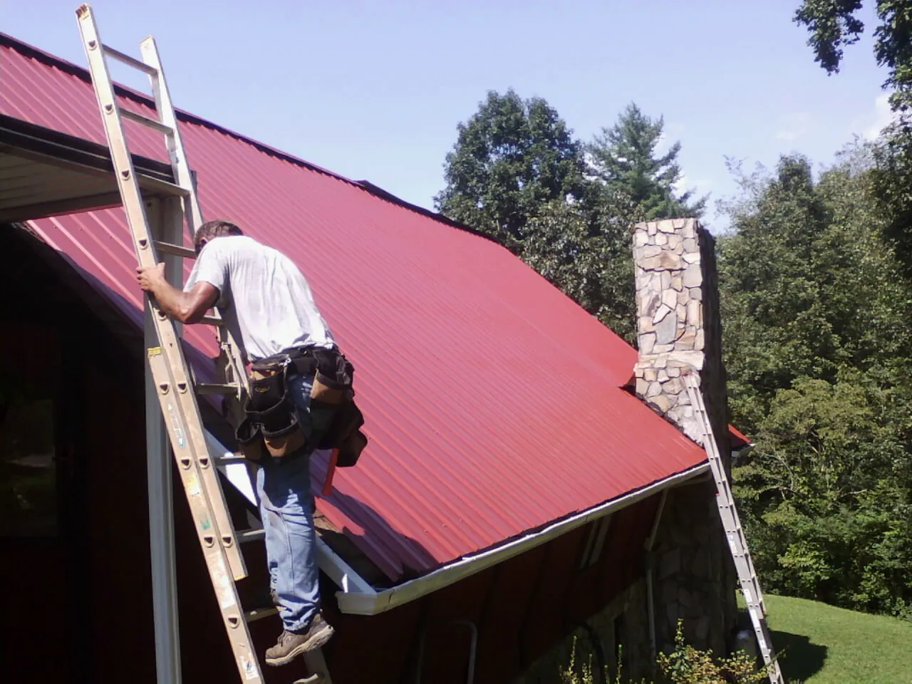 Roofer on a ladder beside a red metal roof and stone chimney — Soffit & Fascia Repair by Roofing Brothers in Pewee Valley